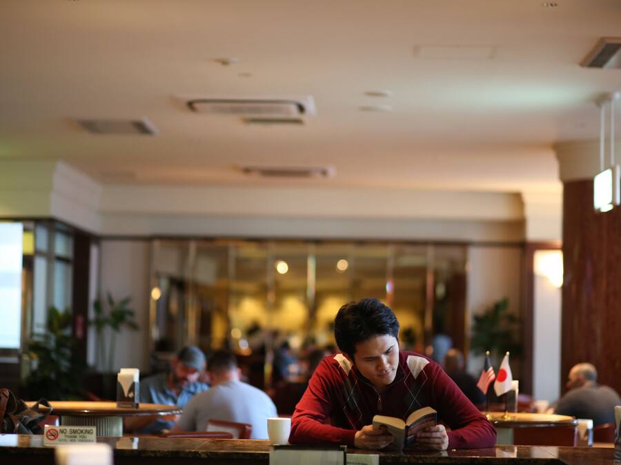 Uma pessoa lendo um livro numa mesa em restaurante, com outros clientes ao fundo.