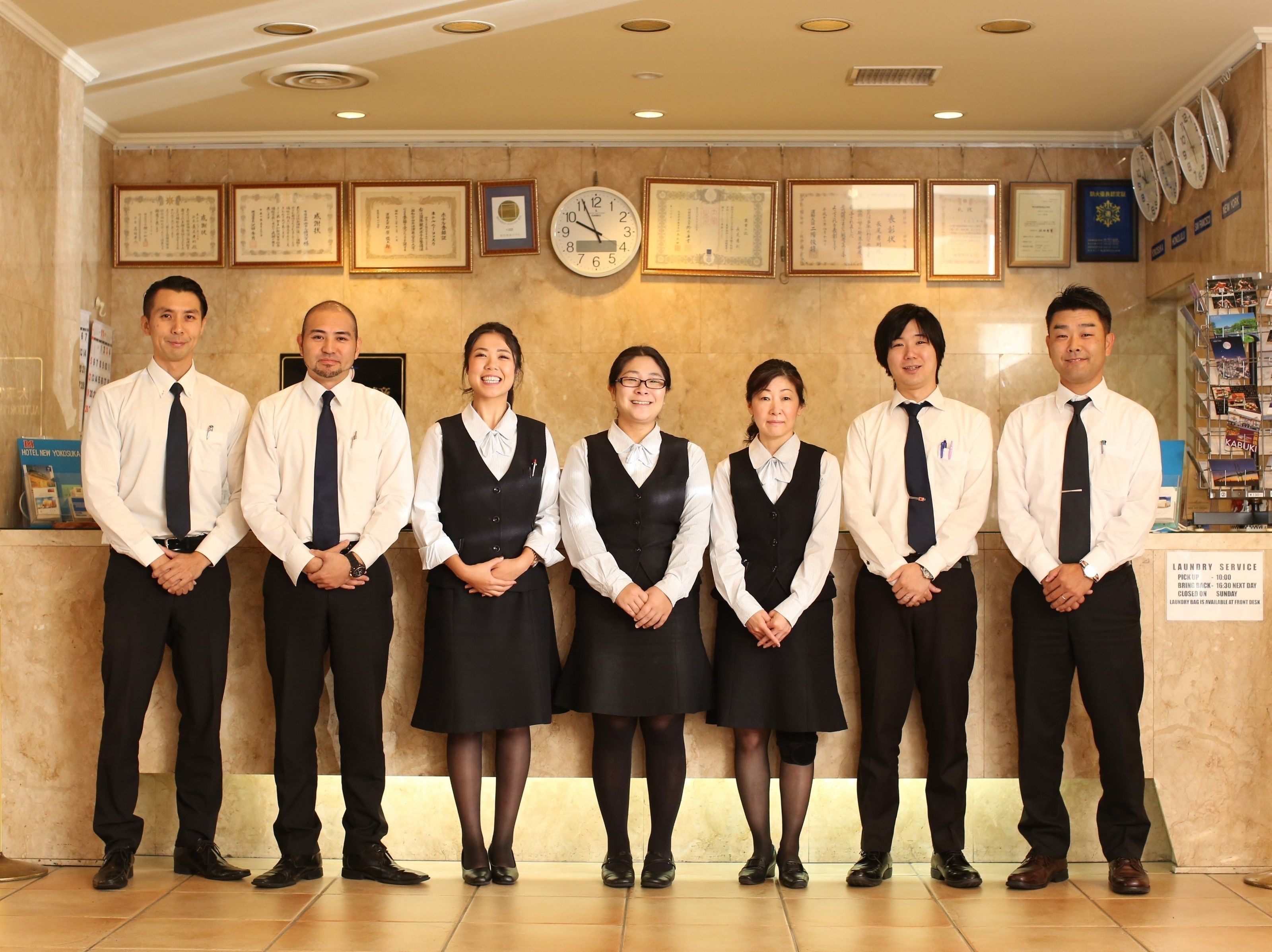 A group of people standing in a reception area with framed certificates on the wall, a clock, and a sign indicating laundry service hours.