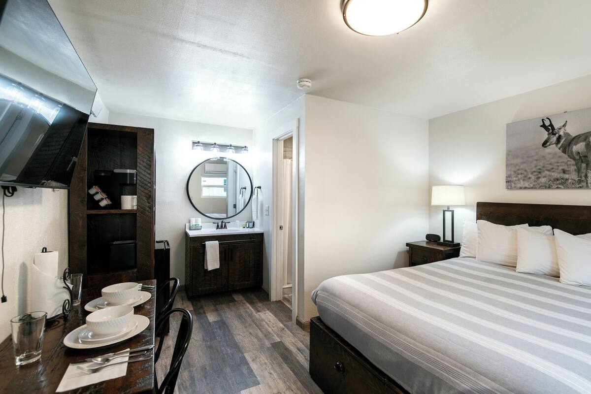 A hotel room with a bed, a mirror, a TV, a sink, and a dining table with plates and glasses.