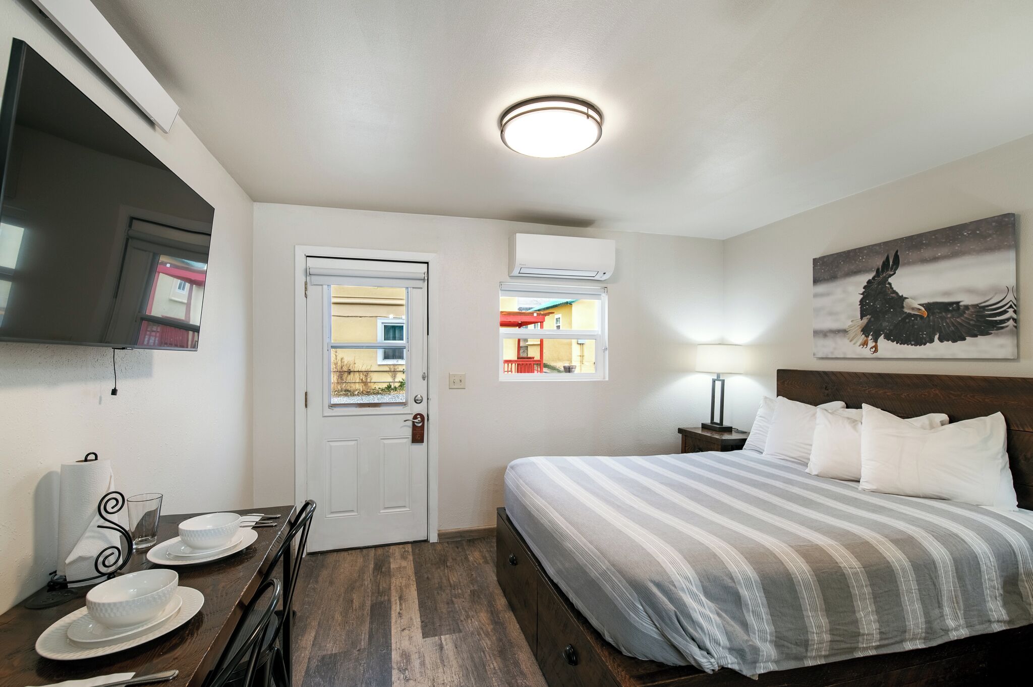 Habitación de hotel con cama, televisor montado en la pared, mesa de comedor con platos y vaso, y ventana con vista a edificios.