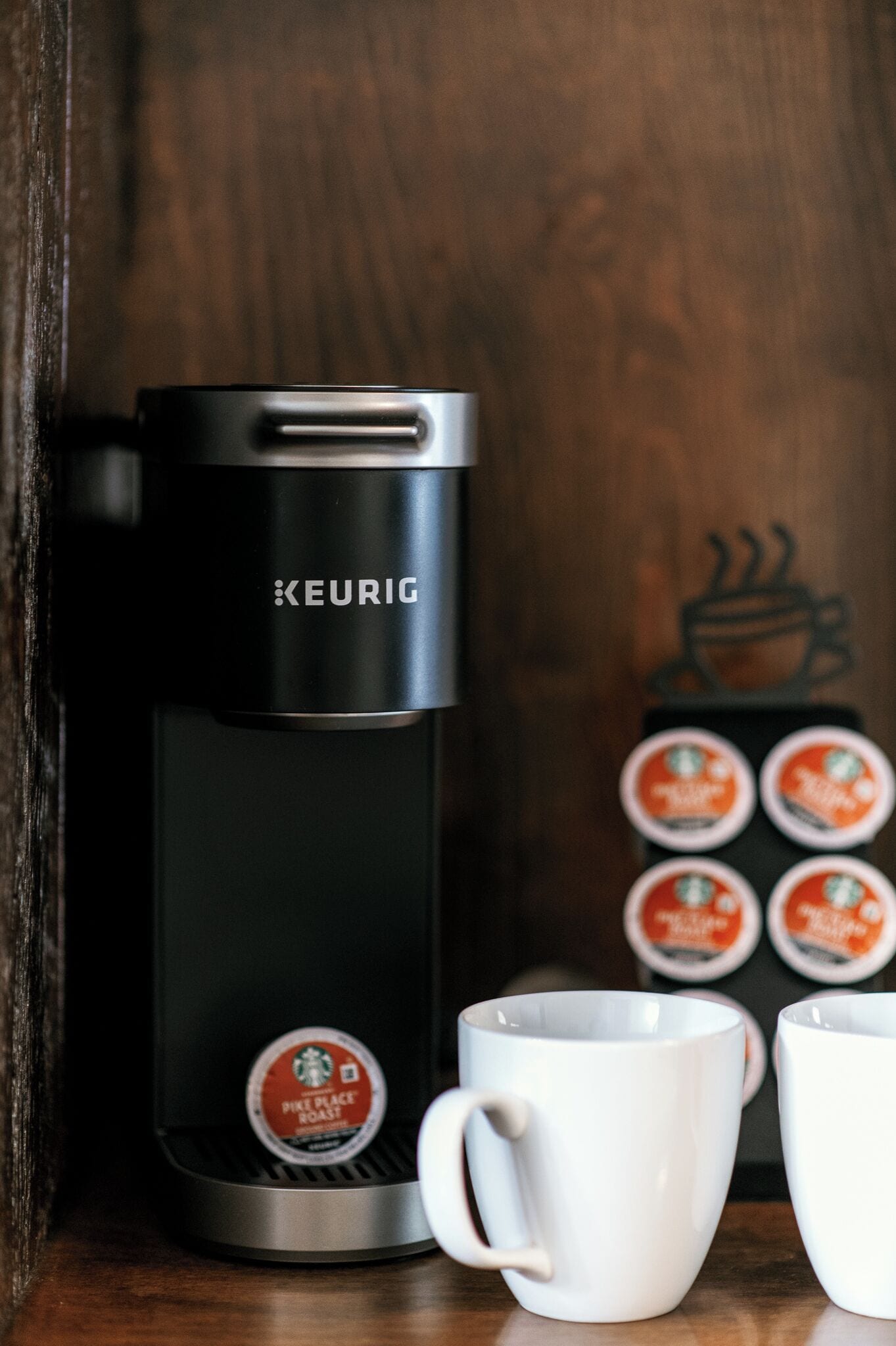 A Keurig coffee maker with a single coffee pod, two white mugs, and a coffee pod holder with multiple pods.