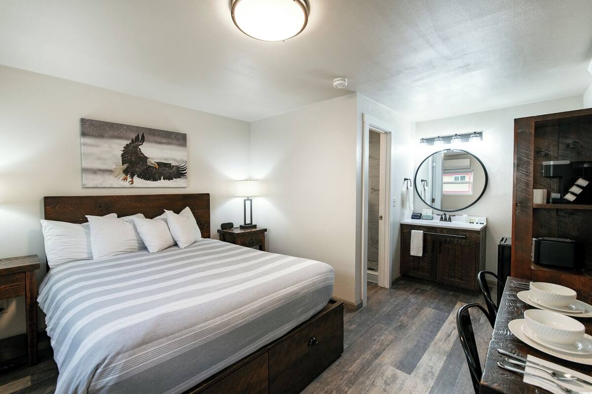 A bedroom with a bed, bedside tables, a nightstand, a mirror, a bathroom with a sink and mirror, and a dining area with chairs and plates.