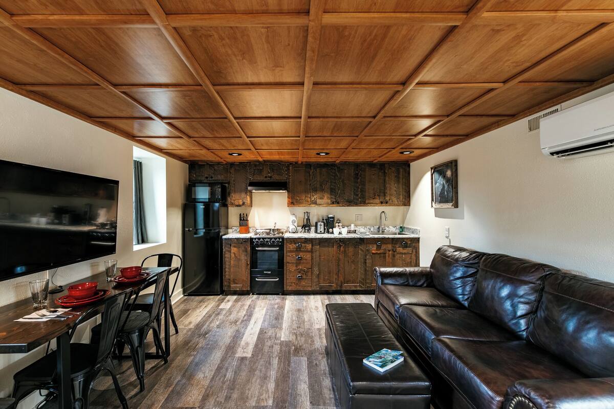 A modern kitchen with wooden ceiling, a dining area with a table and chairs, a leather sofa, and a built-in TV.