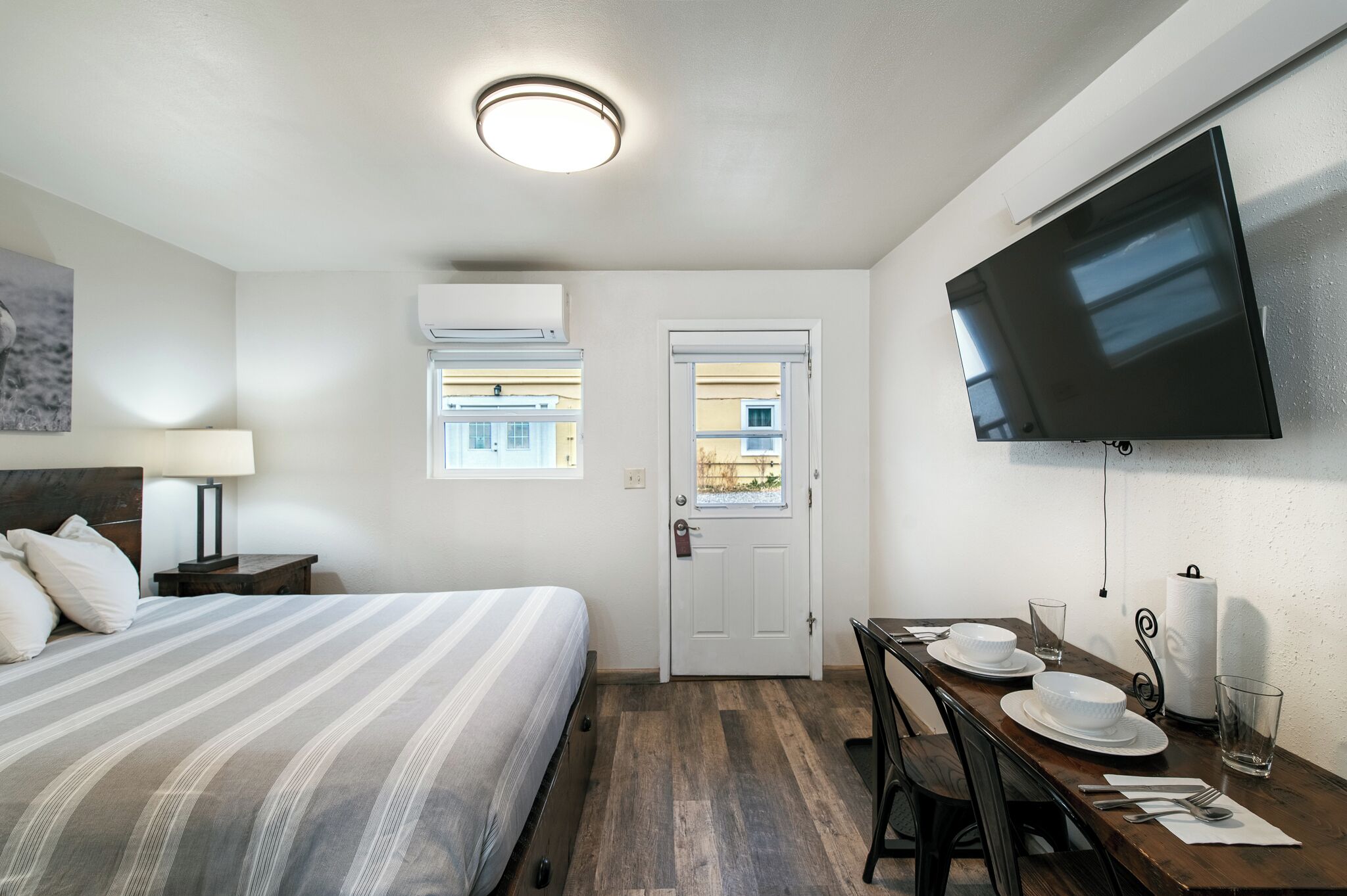 A hotel room with a bed, a dining table set for two, a flat-screen TV mounted on the wall, and an air conditioning unit.