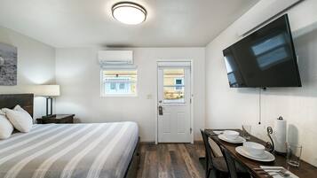 A hotel room with a bed, a dining table set for two, a flat-screen TV mounted on the wall, and an air conditioning unit.