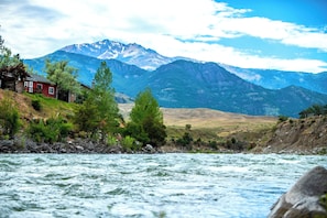 A river with a house on a hillside, mountains in the background, and a clear sky.