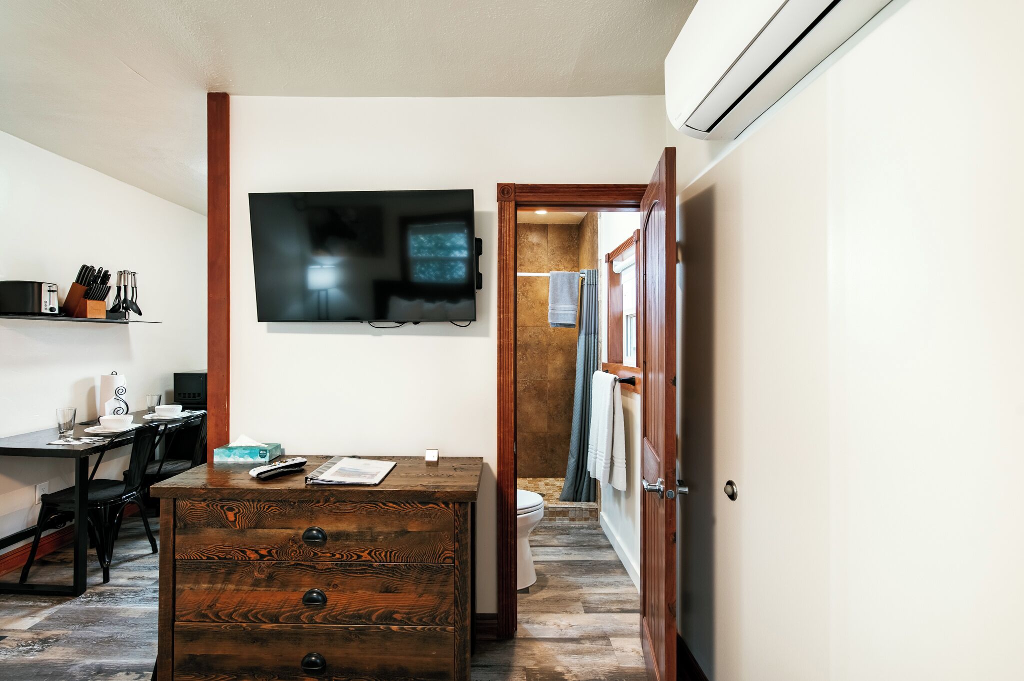 A room with a wooden dresser, a flat-screen TV mounted on the wall, a dining table with chairs, and a bathroom visible through an open door.