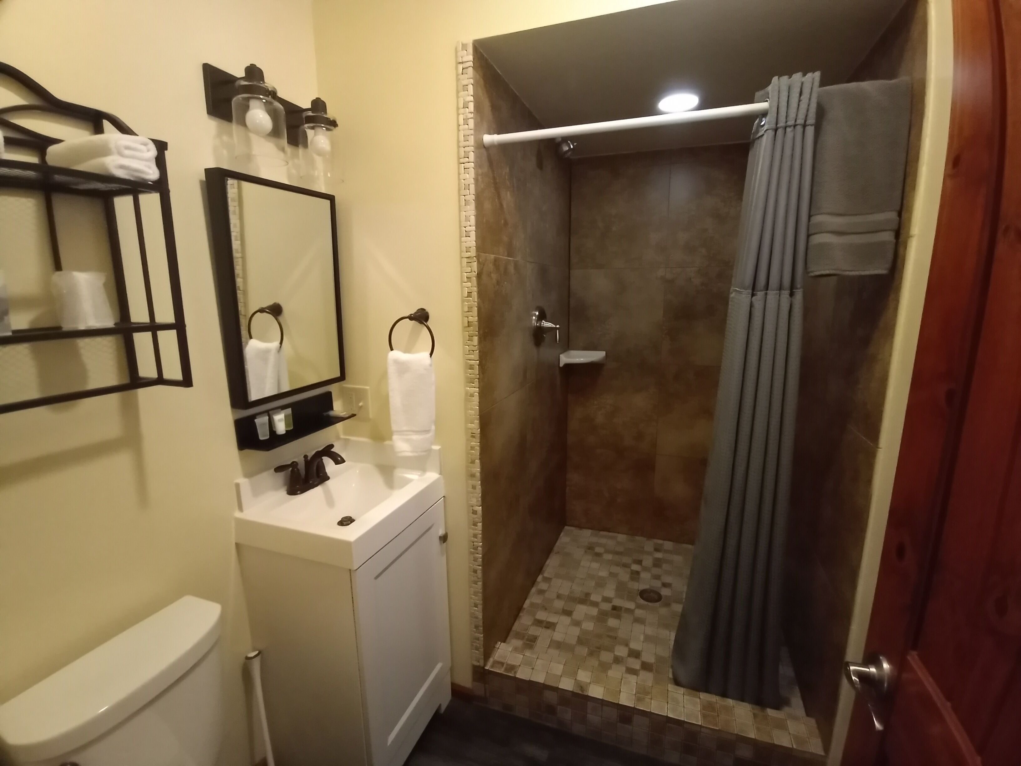 A bathroom with a toilet, sink, mirror, towel rack, shower with a curtain, and a small shelf with toiletries.