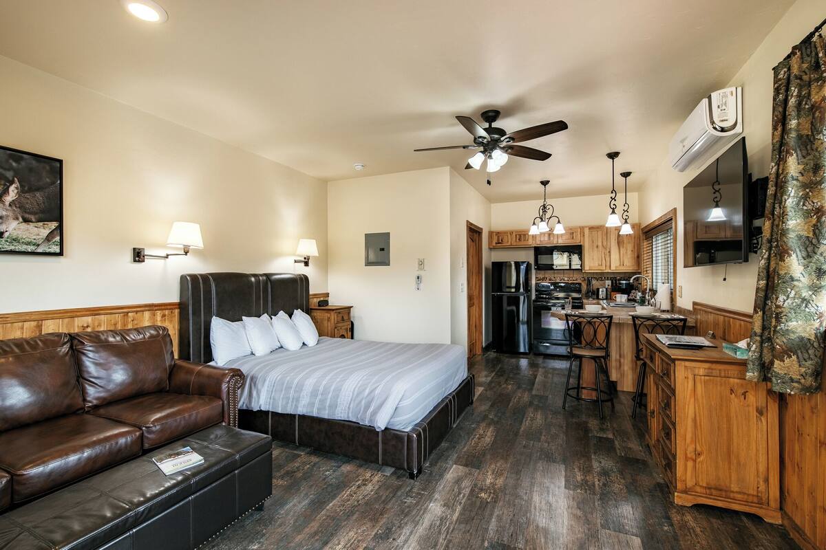 A bedroom with a bed, a leather couch, a ceiling fan, and a kitchenette in the background.