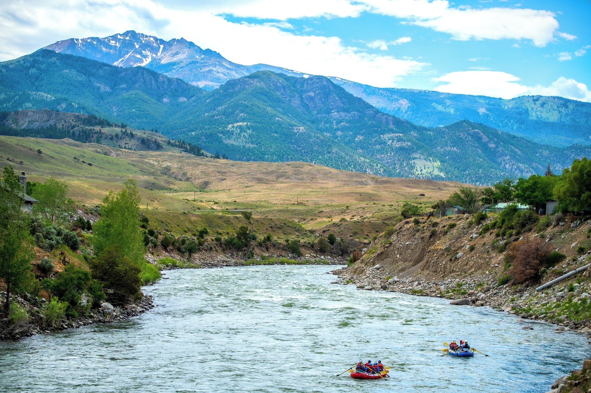 A river with a mountainous backdrop, a raft with two people, and a small building on the left bank.