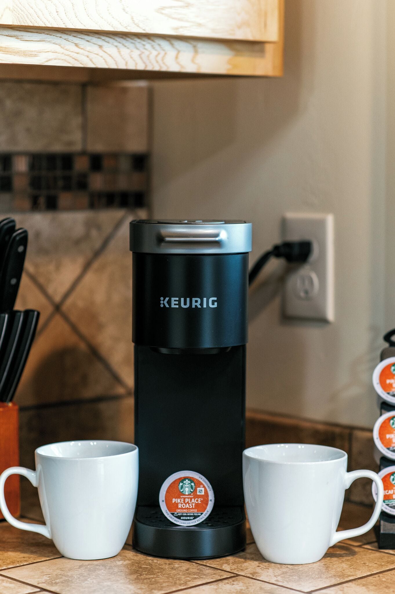Keurig-kahvinkeitin, jossa on kaksi valkoista mukea. Toisessa on Pike Place Roast -merkintä, ja taustalla näkyy keittiö, jonka seinässä on laattapinta.