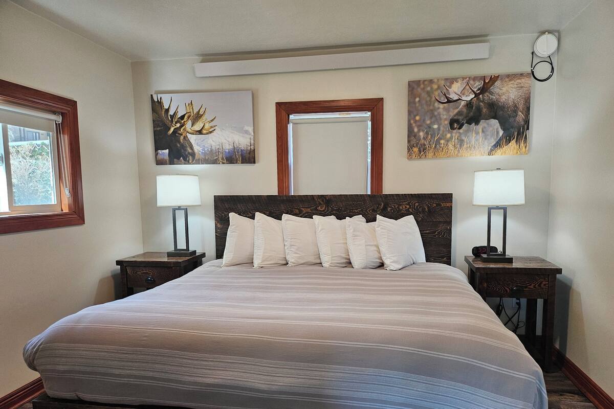 A neatly made bed with a headboard, two bedside tables with lamps, and two wall-mounted paintings of moose.