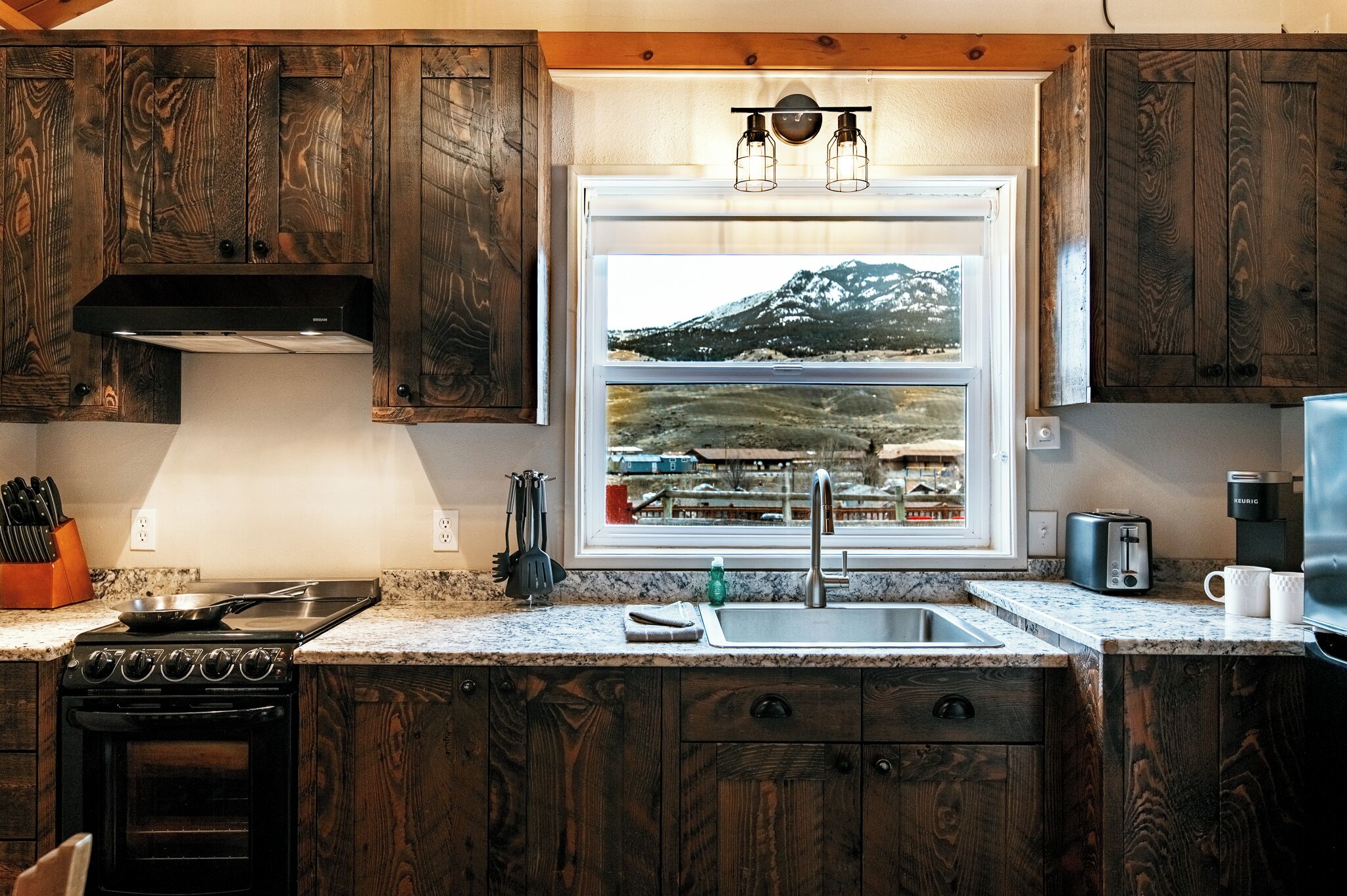 A kitchen with wooden cabinets, a stove, a sink, and a window with a view of a mountain.