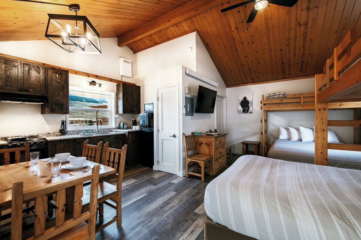 A compact room with a wooden ceiling, a bunk bed, a dining table with chairs, a kitchen area, and a television.