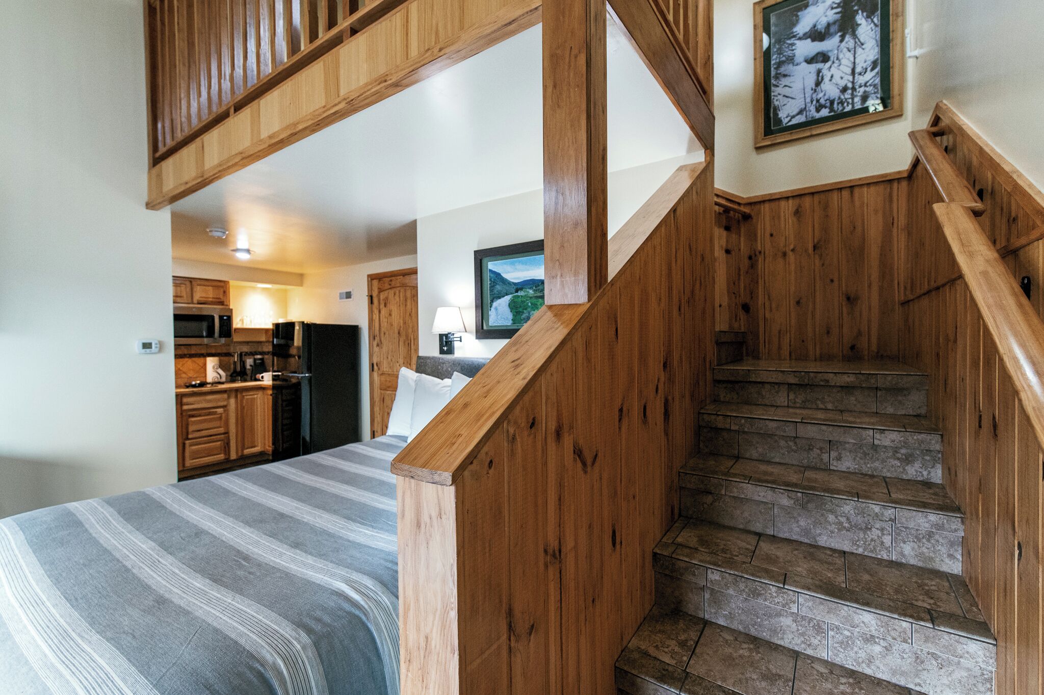 Une chambre avec un lit, un escalier en bois, un coin cuisine et un tableau encadré au mur.