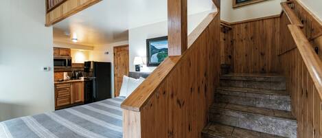 Une chambre avec un lit, un escalier en bois, un coin cuisine et un tableau encadré au mur.