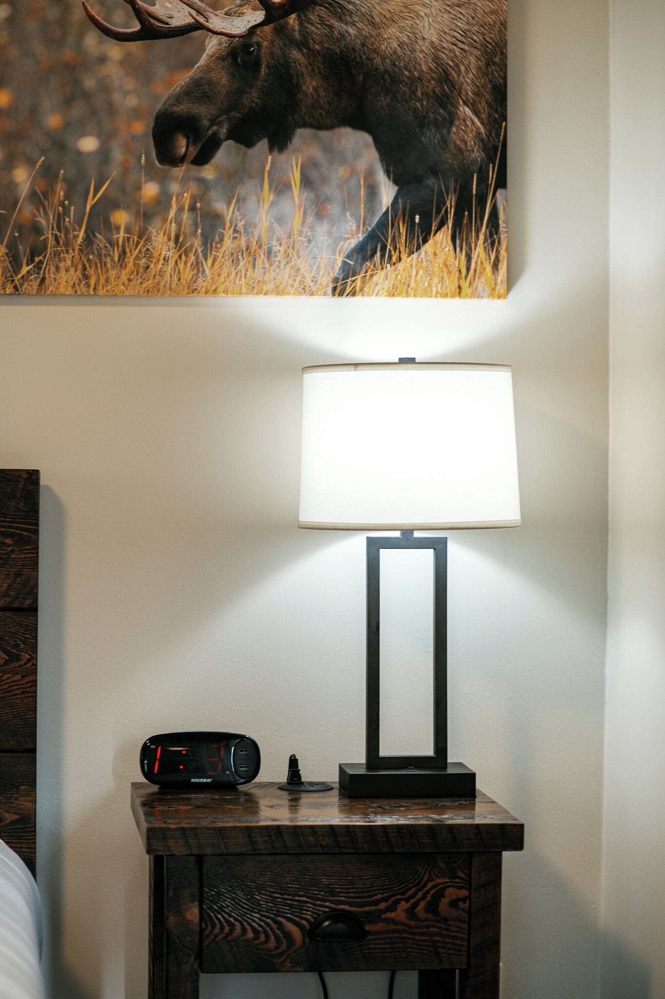 A nightstand with a lamp, a clock, and a moose painting on the wall.