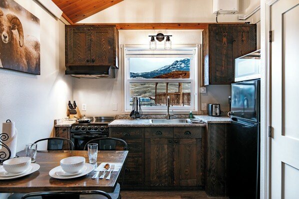 A compact kitchen with wooden cabinets, a stove, a sink, and a window with a mountain view. - Yellowstone Riverside Cottages (Gardiner)