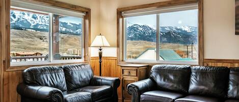 A room with two leather sofas, a wooden desk, and large windows with mountain views.