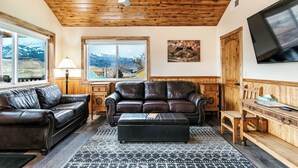 Una sala de estar con sofá de cuero, reposapiés, mesa de centro de madera y televisor montado en la pared.