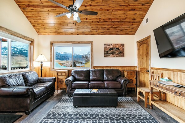 A living room with a leather sofa, ottoman, wooden coffee table, and a TV mounted on the wall. - Yellowstone Riverside Cottages (Gardiner)