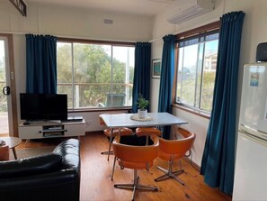 Ein Zimmer mit Fernseher, einem Esstisch mit orangefarbenen Stühlen, einer schwarzen Ledersofa und einem Kühlschrank.