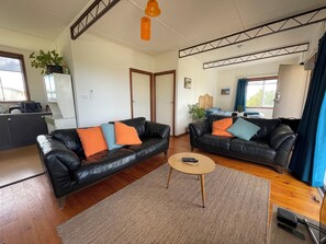 A living room with two black leather sofas, a small round table, and a kitchen area visible in the background. - Cosy retro weekender with far-reaching views (Goolwa Beach)