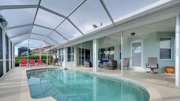 Una piscina interior con paredes de vidrio transparente, sillas de descanso rojas y azules, y un área de patio cubierta.
