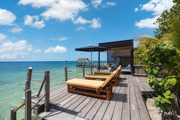 A wooden deck with lounge chairs and a covered seating area overlooking a clear blue sea.