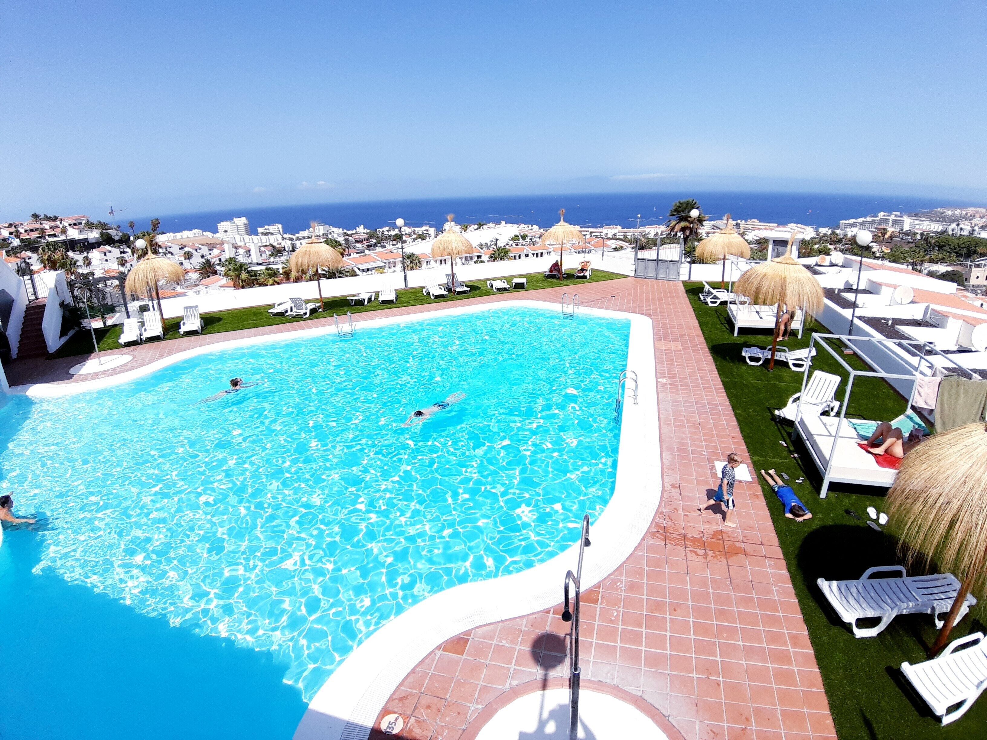 Una piscina amb aigua clara i blava, envoltada de gandules i ombrel·les blanques, amb vista al mar i edificis al fons.