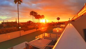 Un balcón con vistas a un atardecer, palmeras y un edificio al fondo.