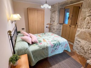 A bedroom with a bed, a nightstand, a lamp, a wardrobe, and a stone wall.