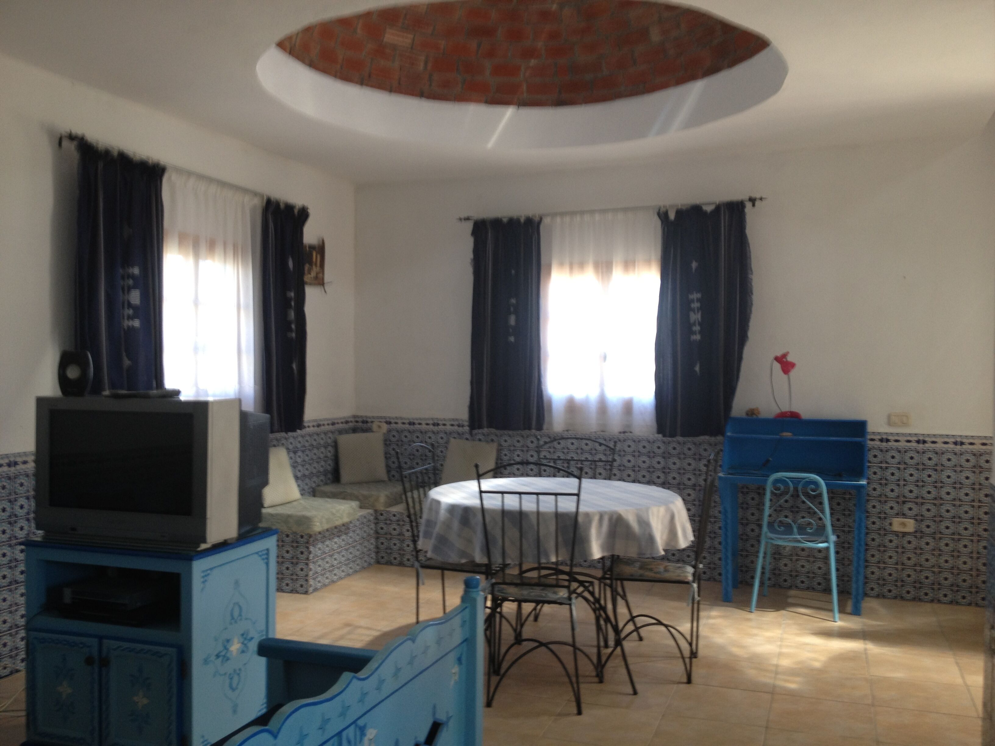 Une pièce avec un téléviseur posé sur un meuble, une table ronde entourée de chaises, un meuble bleu et un mur orné d’un motif.