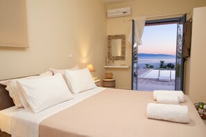 A bedroom with a bed, pillows, a bedside table with a lamp, a mirror, and a view of the sea through an open door. - Trapezaki Villas, Seafront Accommodation (Karavados)