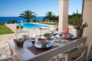 A dining table set with a salad, bowls, and glasses, overlooking a swimming pool and the sea. - Trapezaki Villas, Seafront Accommodation (Karavados)