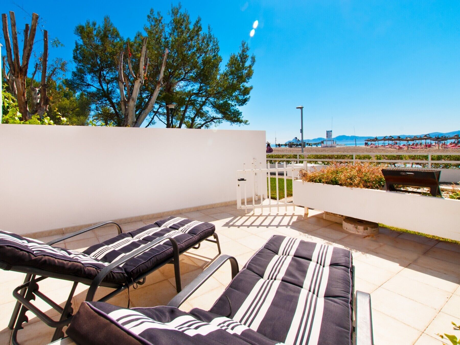 A rooftop terrace with two sun loungers, a white wall, and a view of a beach with umbrellas.