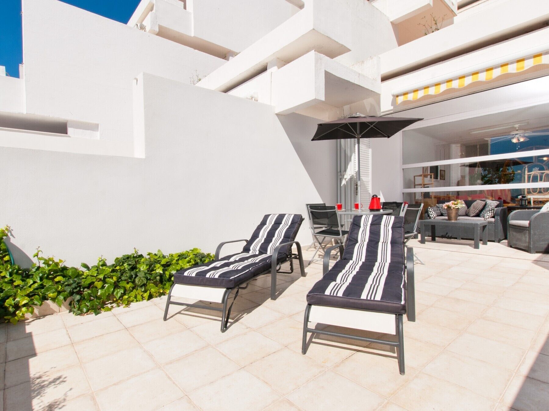 Ein sonniger Außenbereich mit Sitzgelegenheiten, darunter zwei Lounge-Stühle und ein Tisch, umgeben von einer weißen Mauer und einem kleinen Garten.