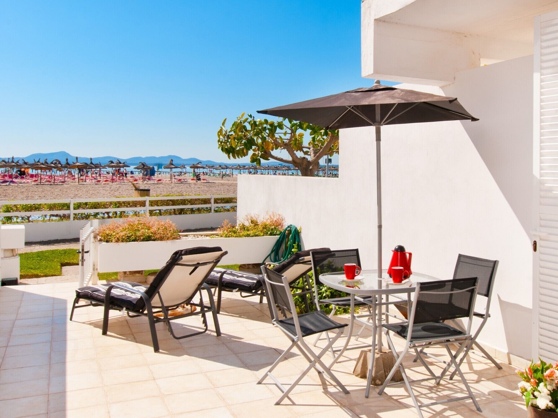 Une terrasse extérieure avec une table et des chaises, un grand parasol, et une vue sur la plage et les montagnes en arrière-plan.