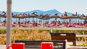 A beachside view with thatched umbrellas and lounge chairs, a mountain in the background, and a table with red cups. - A. Oiza Sand Castle 24, Apt. 24 Edificio Canopus (Alcúdia)