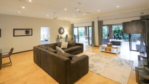 Una sala de estar moderna con un sofá modular, una mesa de centro y un mueble para televisión. Hay ventiladores de techo, un ventanal con cortinas y vistas al exterior.