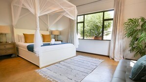 Un dormitorio con cama con dosel, una ventana grande con cortinas, una mesita de noche con lámpara y una planta en maceta.