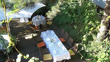 Een terras met tafels voor twee personen, omgeven door groen en bomen.