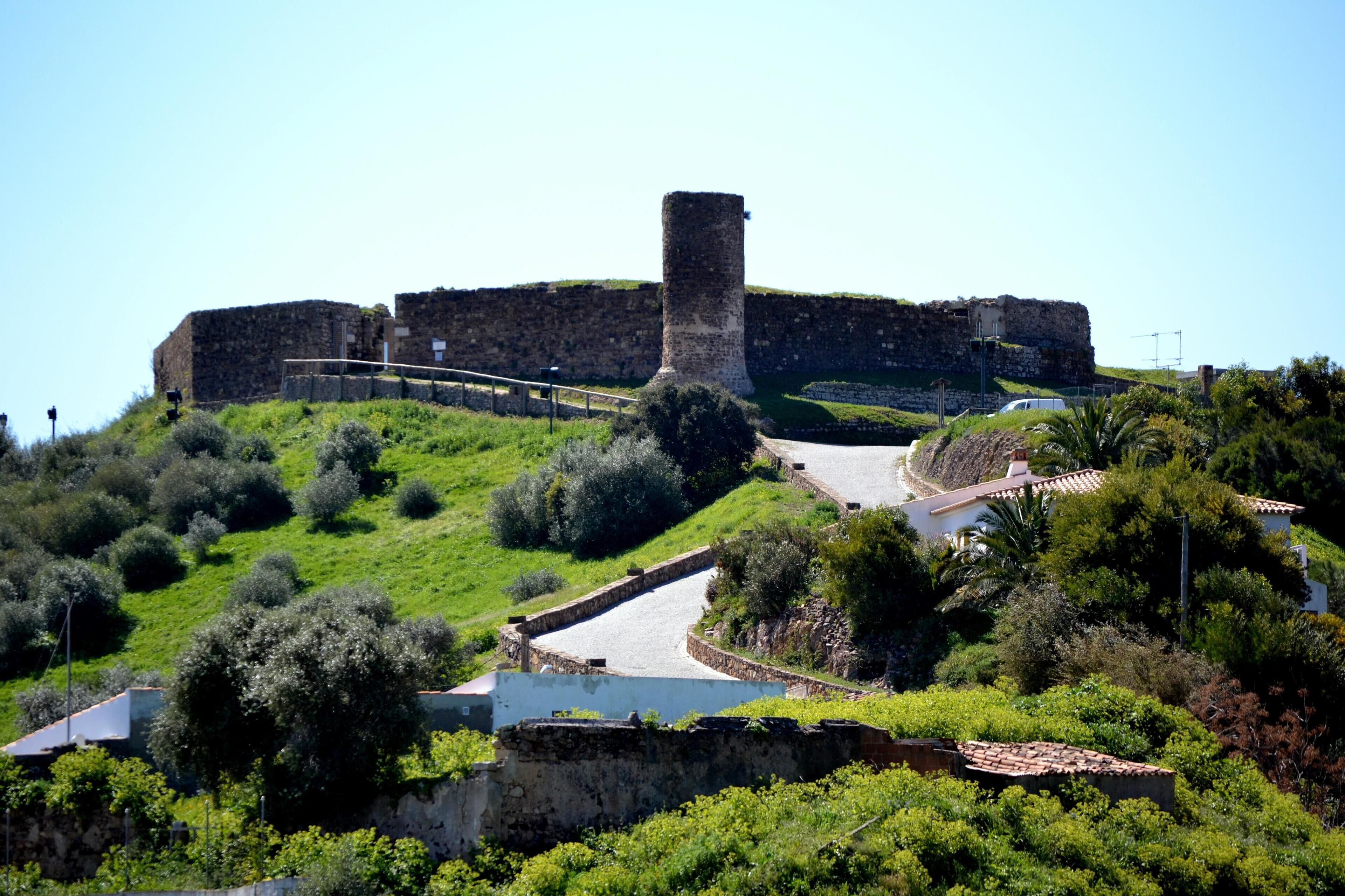 Un’antica fortezza con una torre imponente, circondata da vegetazione rigogliosa e un viale lastricato che conduce a un edificio bianco.