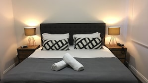 Ein ordentlich bezogenes Bett mit dunklem Kopfteil, zwei Nachttischlampen und zwei aufgerollte Handtücher auf dem Bett.