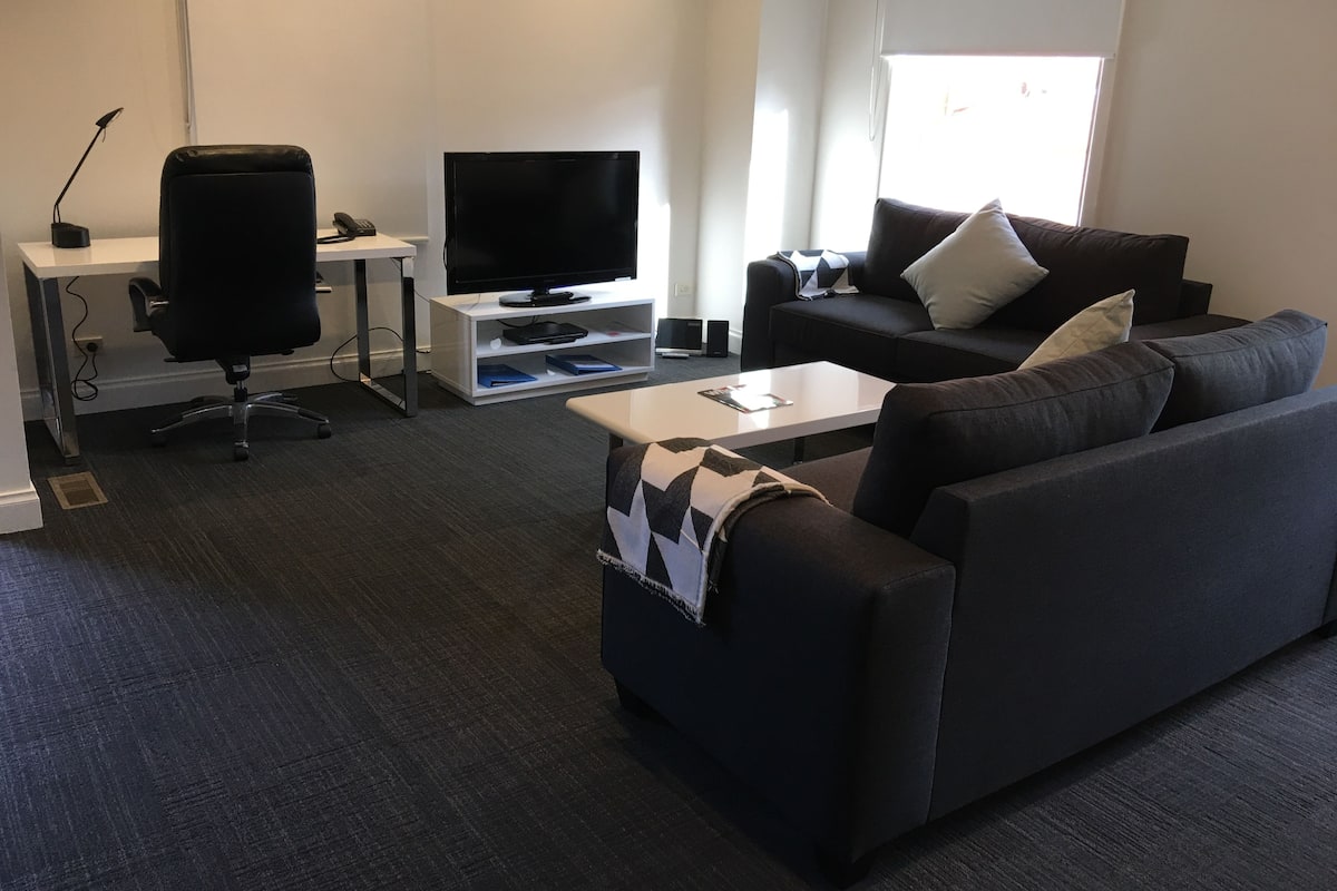 A modern living room with a black leather office chair, a white TV stand, a black sofa, and a coffee table.