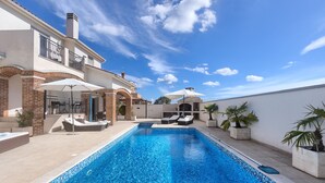 Une piscine entourée de chaises longues et de parasols, bordée d’un bâtiment à deux étages doté d’un toit en tuiles et de détails en briques.