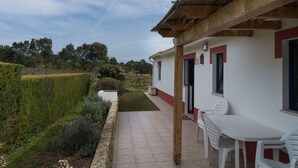 Eine Terrasse mit weißem Tisch und Stühlen, einem gepflasterten Weg und einem Garten mit Hecke und kleinem Baum.