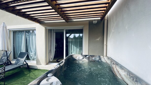 Ein Whirlpool mit einer hölzernen Pergola darüber, ein Terrassenbereich mit Liegestühlen und eine Glastür, die zu einem Innenbereich führt.