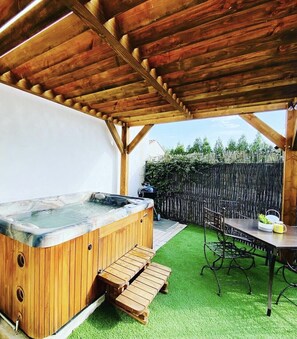 Ein hölzernes Whirlpool unter einer Pergola mit Tisch und Stühlen, umgeben von Kunstgrün und einem Holzzaun.
