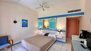 Une chambre d’hôtel avec un lit, un bureau, une chaise, un ventilateur de plafond, un tableau et une porte de salle de bain.